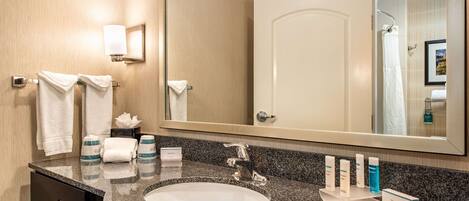 A hotel bathroom with a granite countertop sink, a large mirror, and toiletries on the counter.
