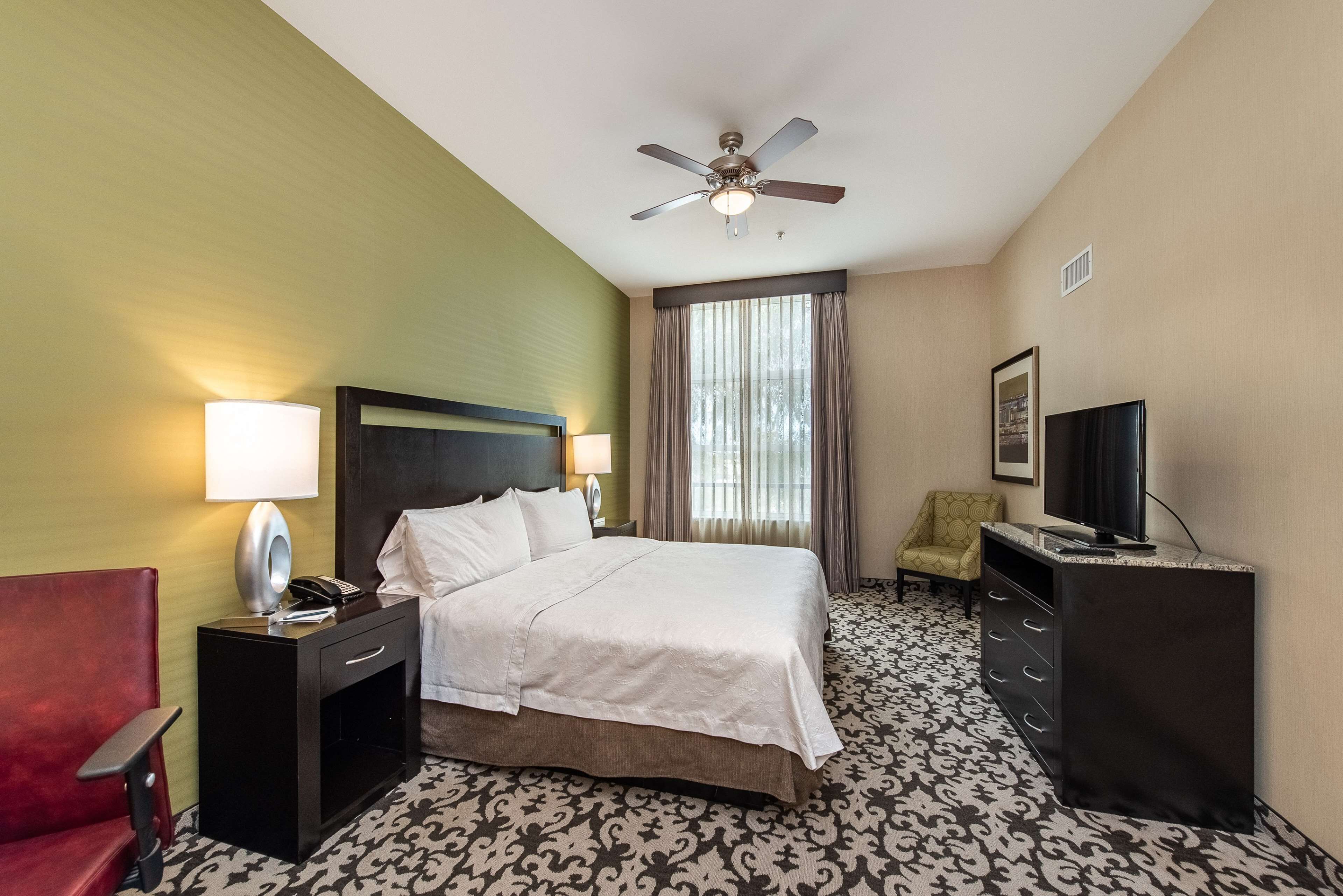 A hotel room with a bed, a desk with a lamp, a chair, a television, and a ceiling fan.