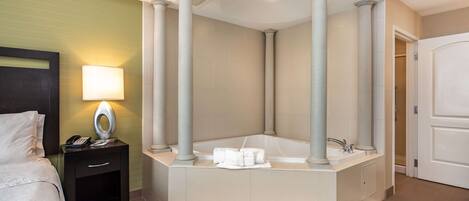 A hotel bathroom with a large bathtub, a bedside table with a lamp, and a bed.