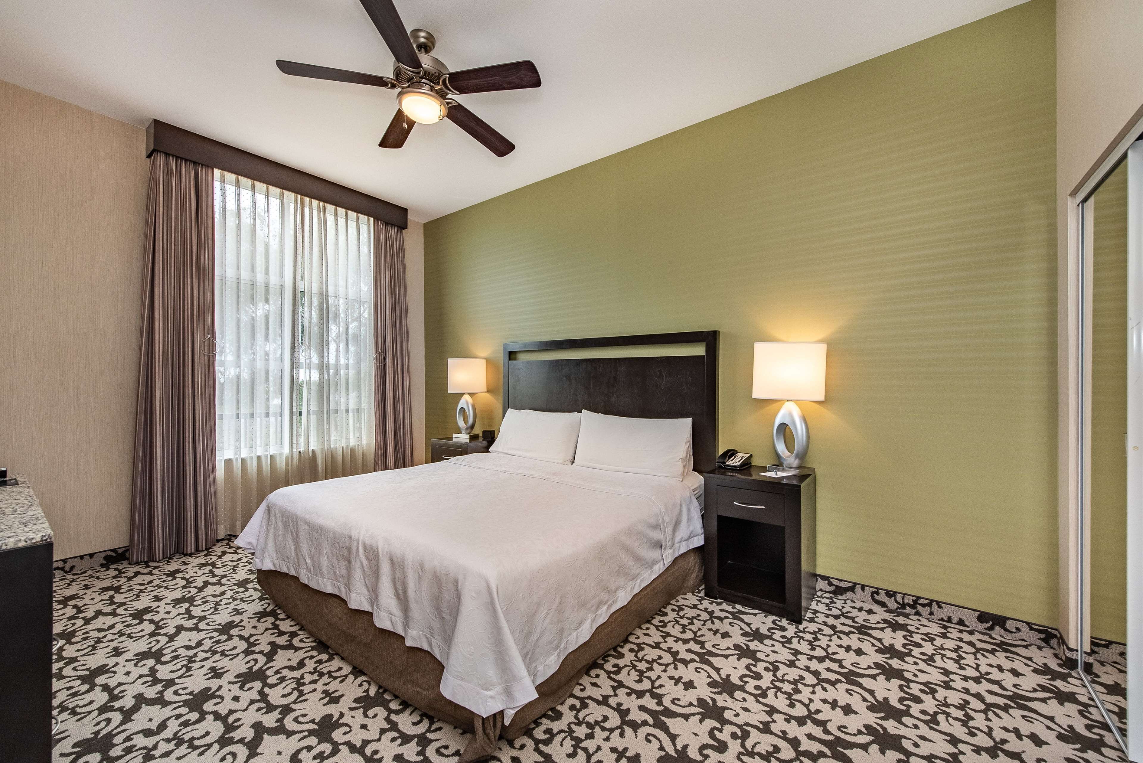 Una camera d'albergo con un letto grande, due comodini con lampade, una ventola a soffitto e un tappeto decorato.