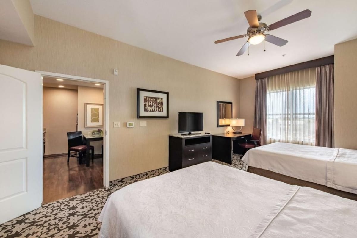 Habitación de hotel con dos camas, un escritorio, televisor, ventilador de techo y una puerta que da a un baño.