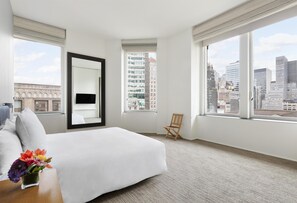 Une chambre d’hôtel dotée d’un grand lit, d’un coin salon avec un fauteuil, d’une petite table ornée d’un vase de fleurs et de grandes fenêtres offrant une vue sur la ville.