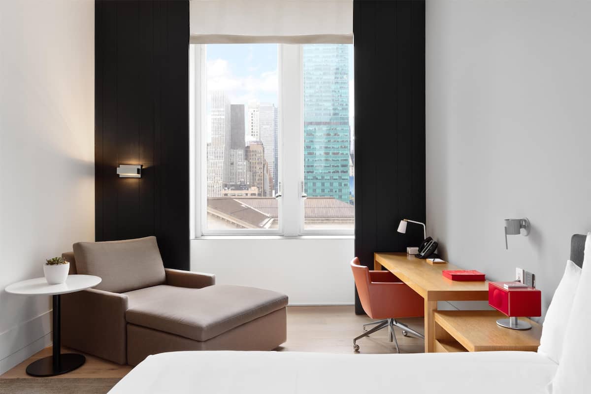 Habitació d'hotel moderna amb un llit, escriptori, cadira, taula petita amb una planta i vista a edificis de la ciutat.