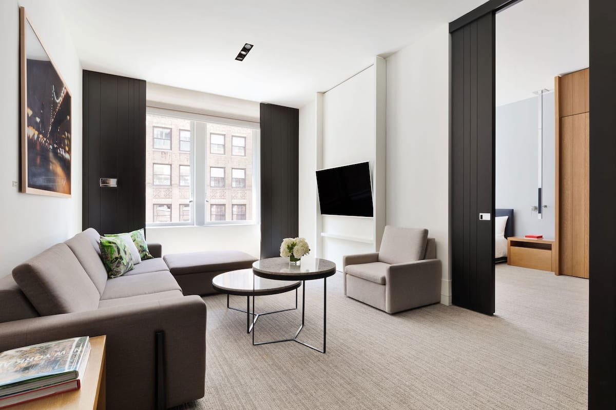 Una habitació d'hotel moderna amb un sofà, una butaca, una taula de centre rodona i un televisor de pantalla plana fixat a la paret.