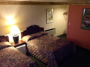 A hotel room with two beds, a nightstand with a lamp, a wall-mounted heater, and a framed picture. - Humboldt Redwoods Inn (Garberville)