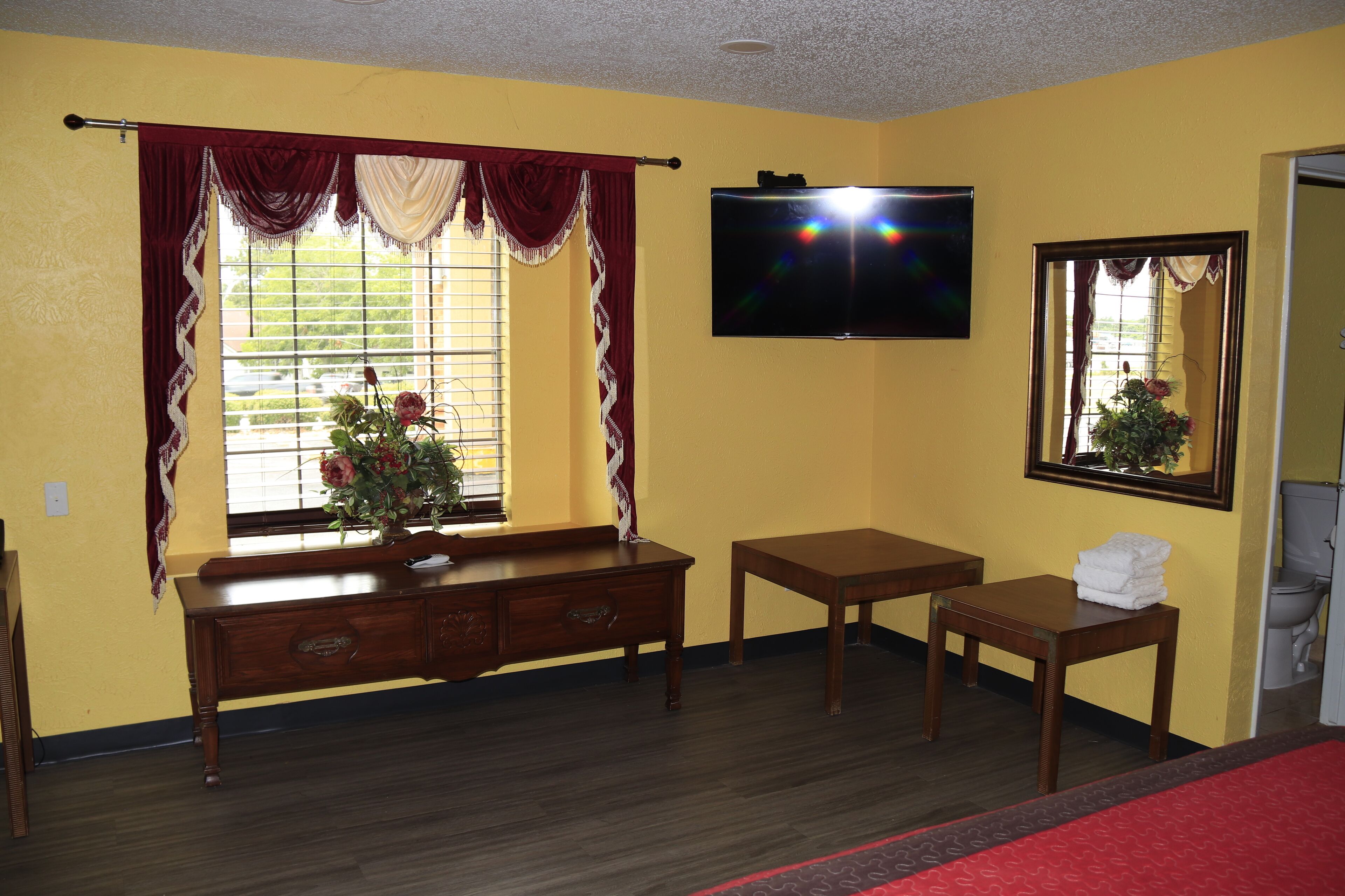 Quarto com escrivaninha de madeira, televisão fixada na parede, janela com cortinas e espelho.