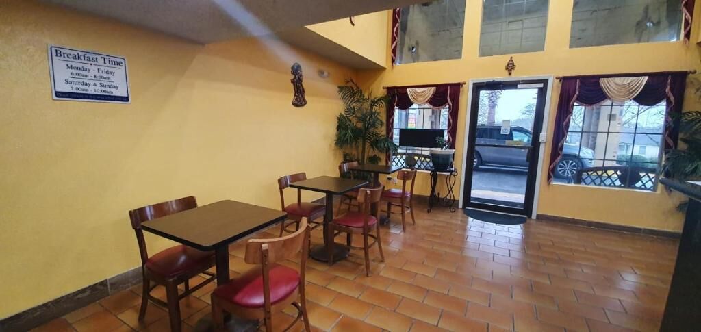 A dining area with tables and chairs, a sign indicating breakfast times, and a view of the outside through a window.
