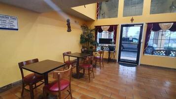 A dining area with tables and chairs, a sign indicating breakfast times, and a view of the outside through a window.