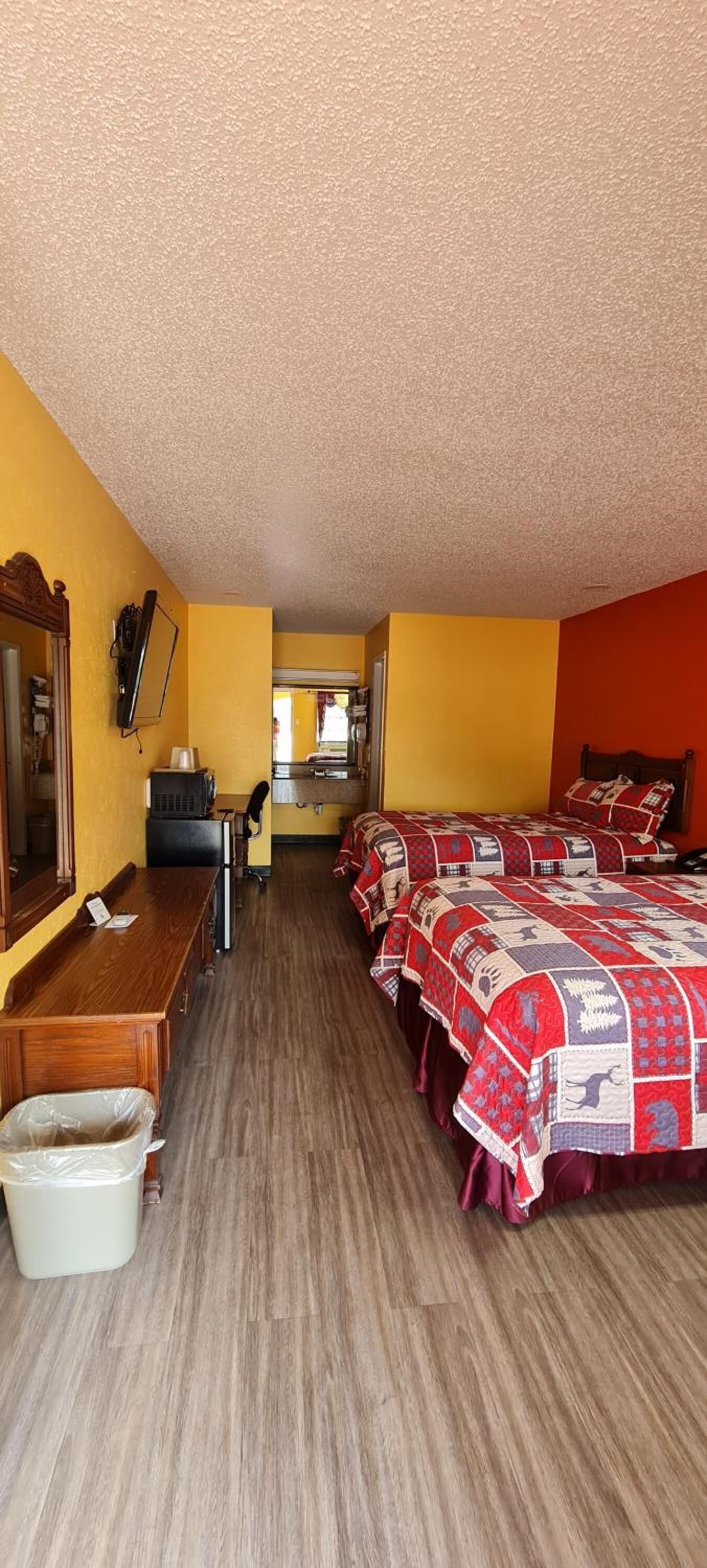 Une pièce avec un bureau en bois, une chaise, un lit recouvert d’une couette rouge et blanche, et un miroir accroché au mur.