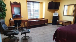 A hotel room with a wooden desk, two black chairs, a bed with a red cover, a television, and a window with blinds. - Chaparral Motel (Hallettsville)