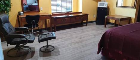 A hotel room with a wooden desk, two black chairs, a bed with a red cover, a television, and a window with blinds.