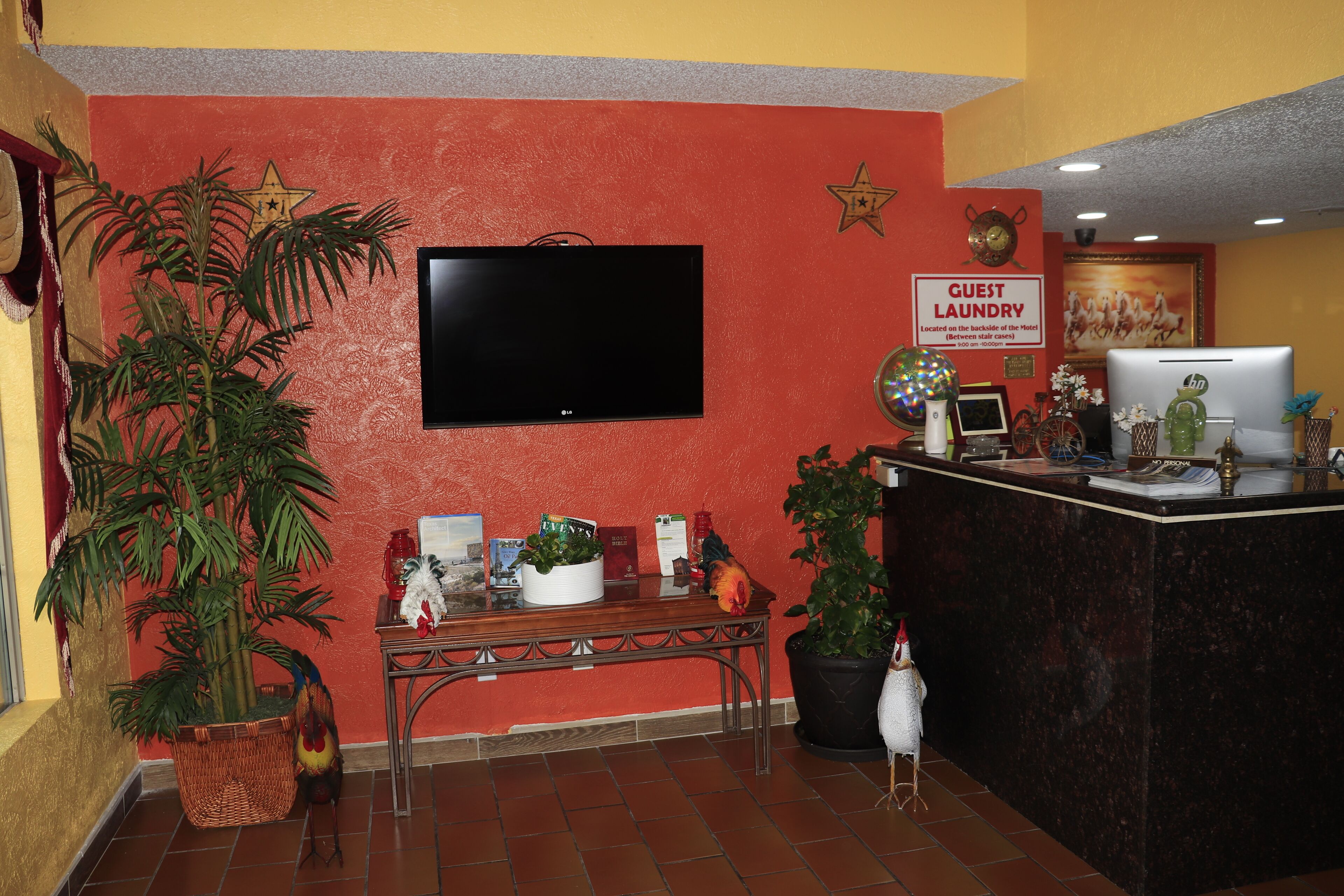 Un hall con televisión, indicación de lavandería para huéspedes, mostrador de recepción y plantas en macetas.
