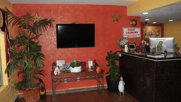 Un hall con televisión, indicación de lavandería para huéspedes, mostrador de recepción y plantas en macetas.