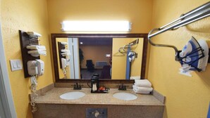 A bathroom with two sinks, a mirror, a hairdryer, and a trash bin. - Chaparral Motel (Hallettsville)