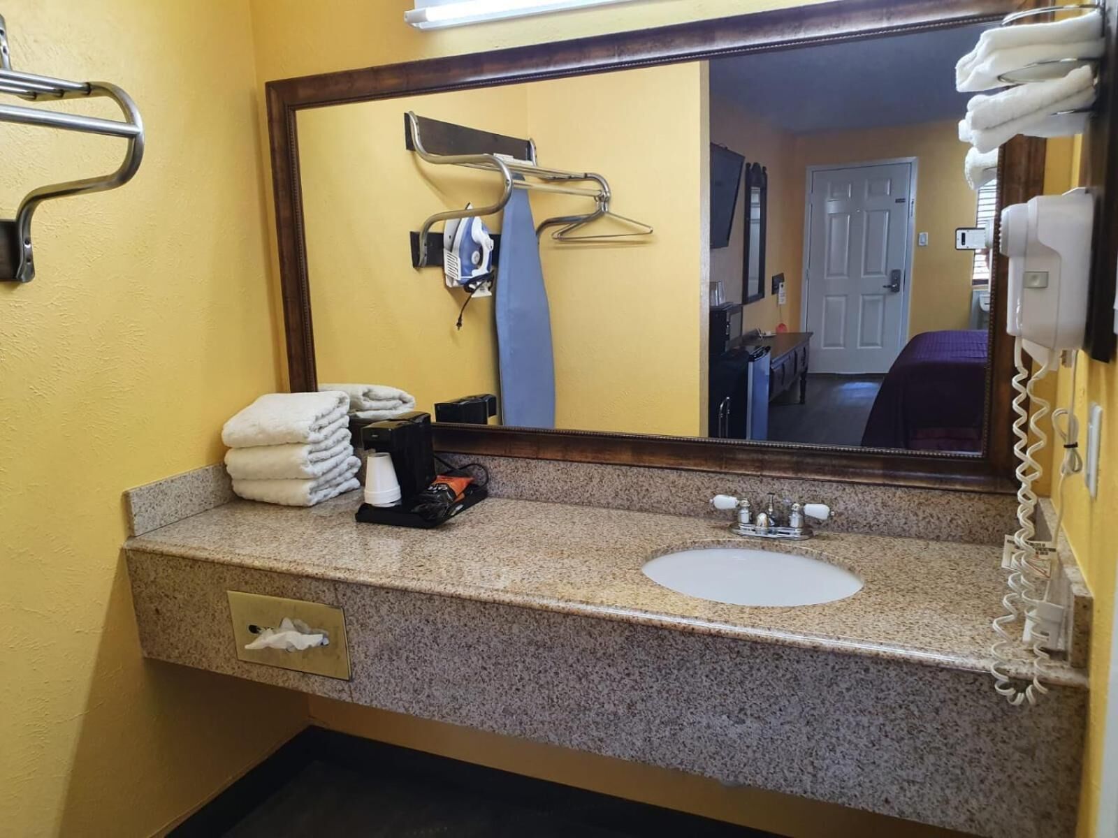 Un baño de hotel con encimera de granito, un lavamanos, un espejo, un perchero para toallas y un teléfono.