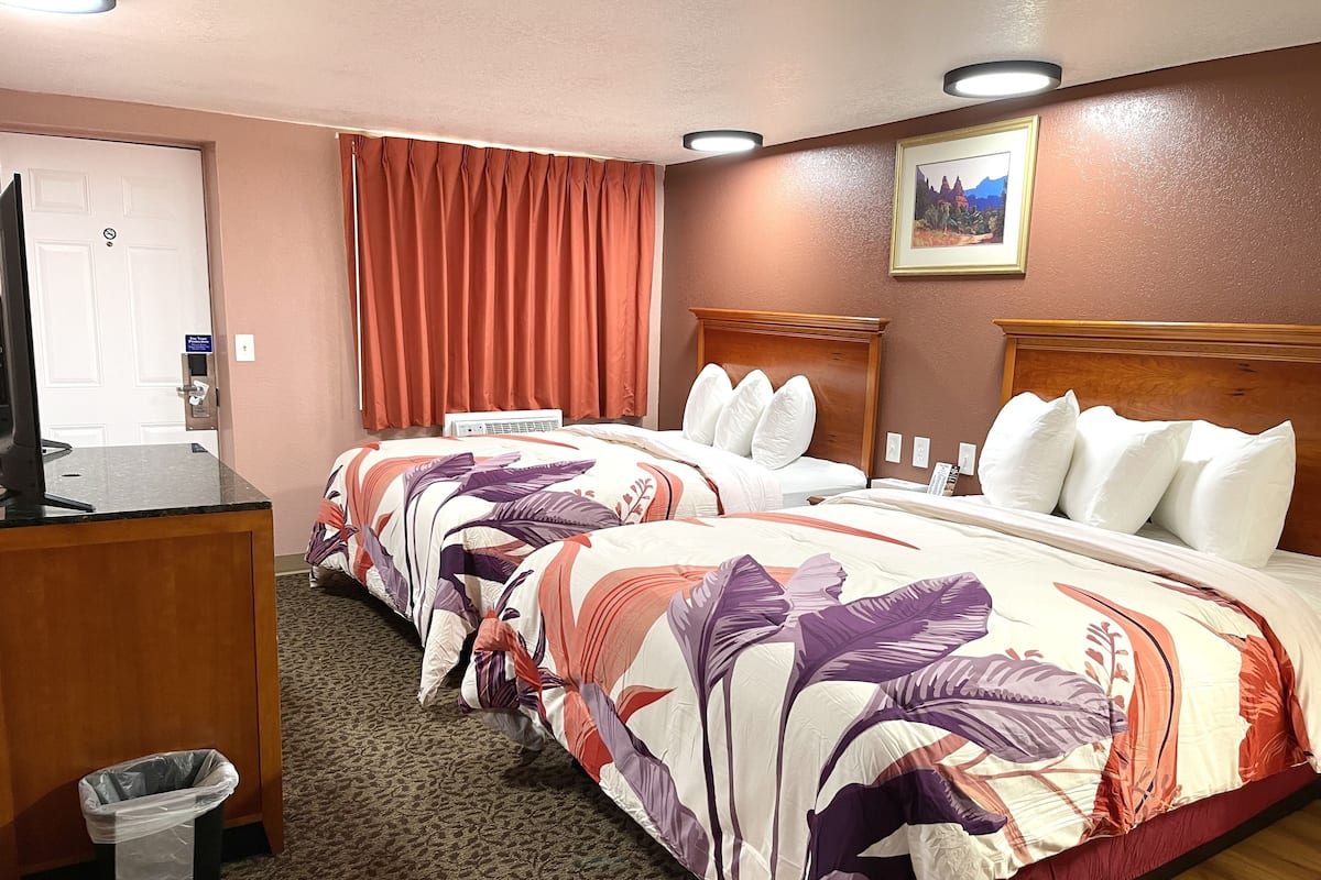 A hotel room with two beds, a desk, a trash bin, and a painting on the wall.