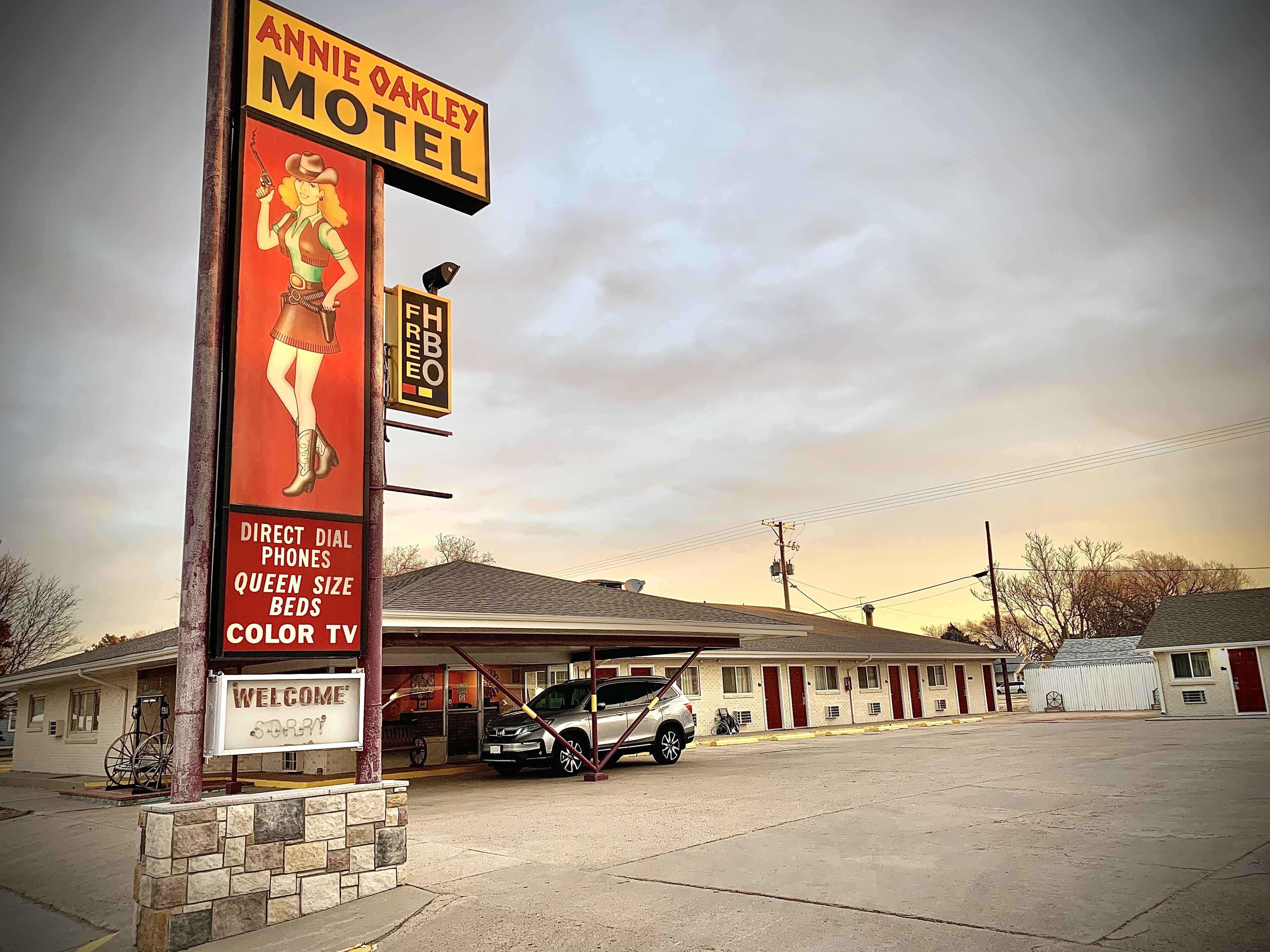 A motel with a large sign featuring a woman in a western outfit, a direct dial phone, queen size beds, and a color TV.