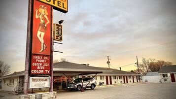 A motel with a large sign featuring a woman in a western outfit, a direct dial phone, queen size beds, and a color TV.