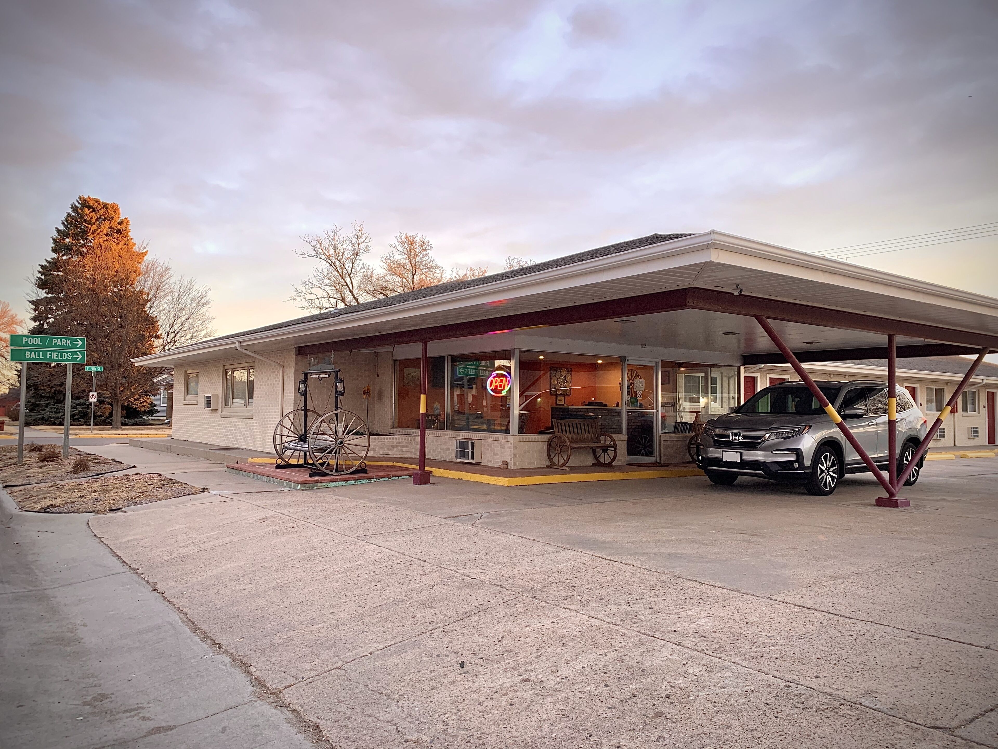 A gas station with a covered parking area, a parked car, and a sign indicating directions to a pool, park, and ball fields.