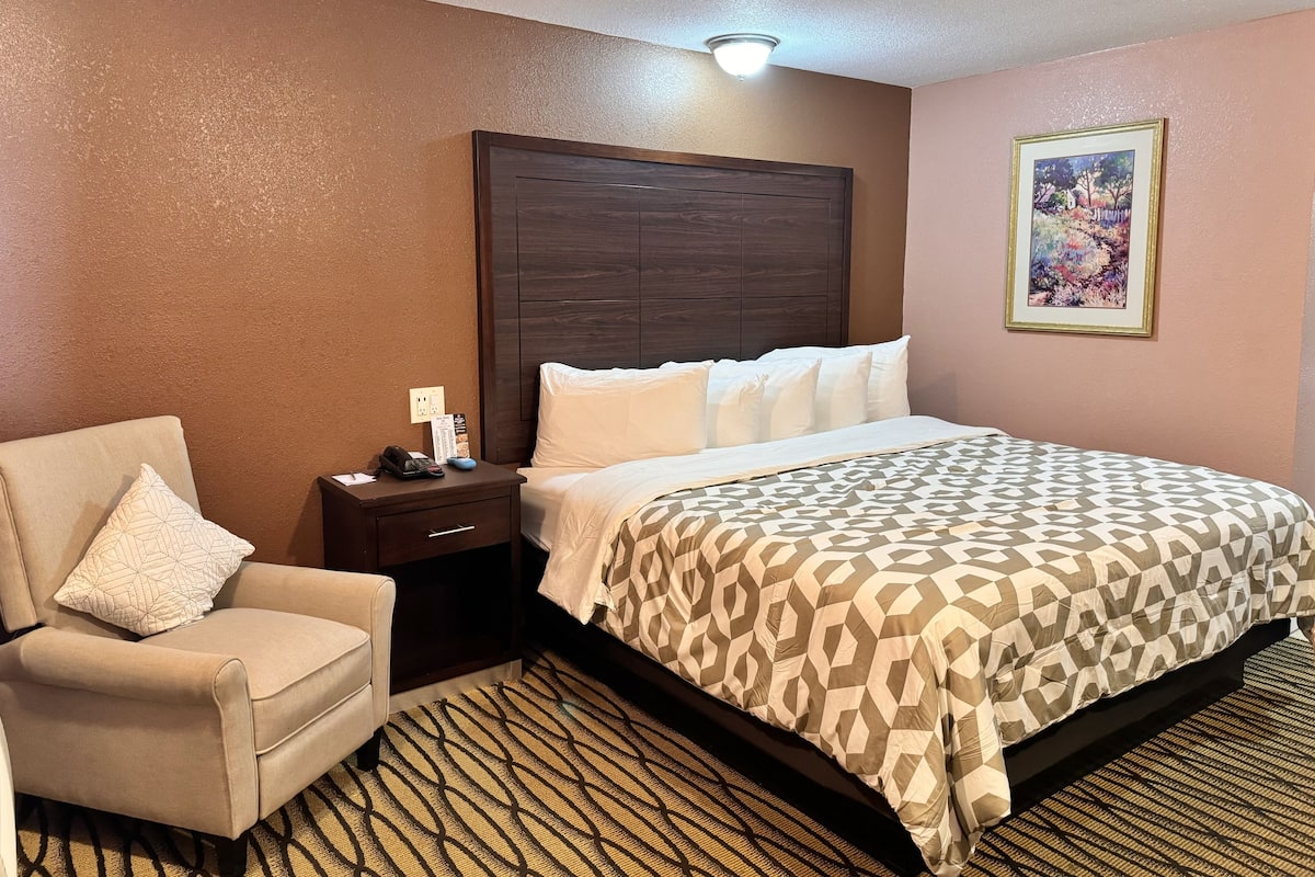 A hotel room with a bed, a chair, a nightstand, and a painting on the wall.