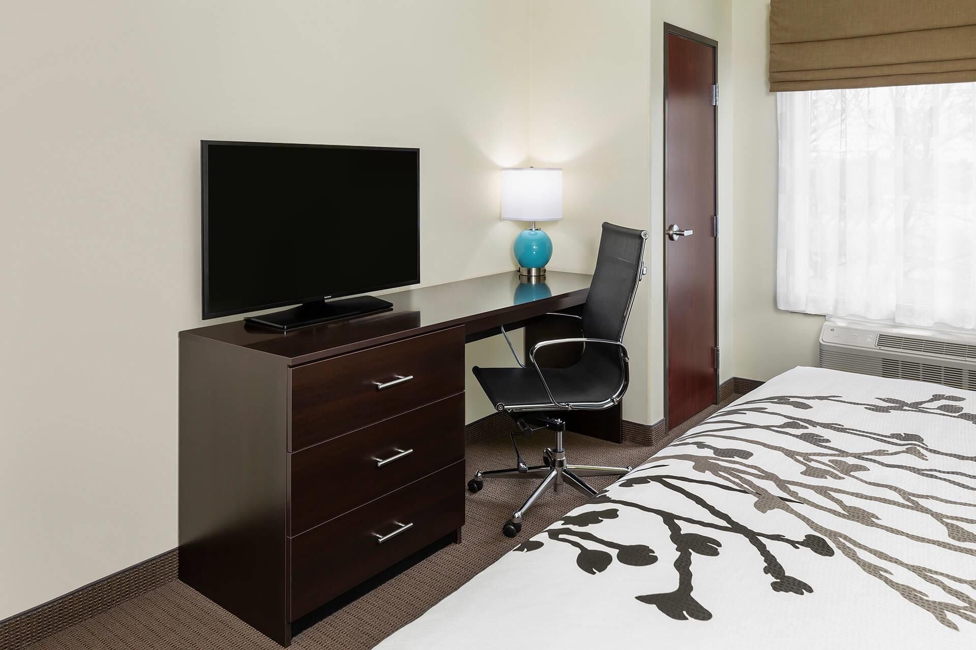 Habitación de hotel con un escritorio de madera oscura, un televisor de pantalla plana, una lámpara azul, una silla de oficina negra, una cama con colcha estampada y una ventana con cortinas blancas.