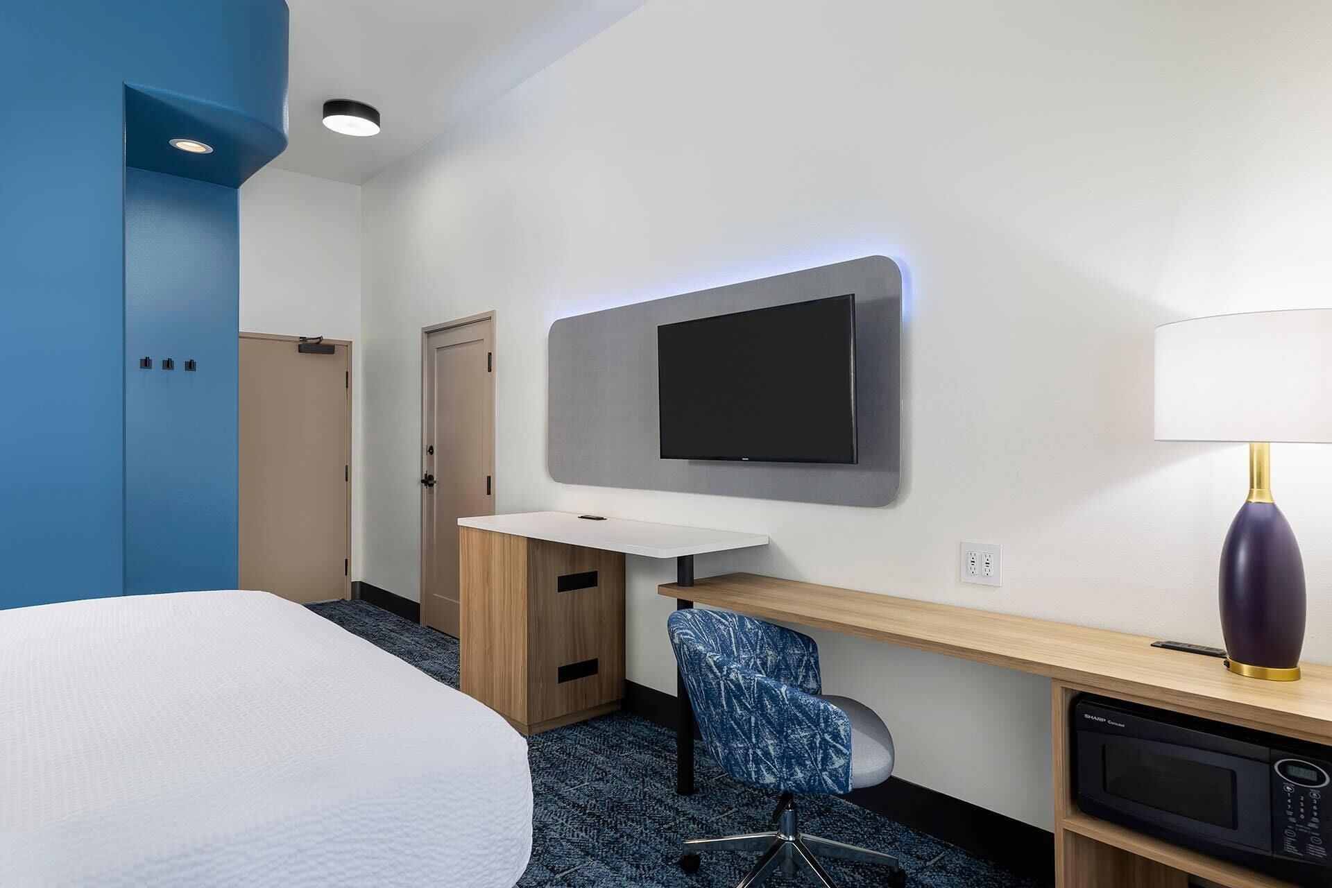 Una habitación de hotel con cama, escritorio con silla, televisor de pantalla plana, lámpara y microondas.