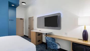 Una habitación de hotel con cama, escritorio con silla, televisor de pantalla plana, lámpara y microondas.