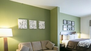A hotel room with a sofa, a small table with a plant, a bed with a white comforter, and wall art. - Sleep Inn & Suites Round Rock - Austin North (Round Rock)