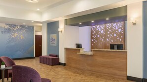 A hotel reception area with a wooden reception desk, a seating area with purple chairs, and a wall-mounted sign for Sleep Inn by Ochoze Hotels. - Sleep Inn & Suites Round Rock - Austin North (Round Rock)