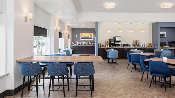 A modern dining area with wooden tables and blue chairs, a kitchen in the background, and a large window providing natural light. - Sleep Inn & Suites Round Rock - Austin North (Round Rock)