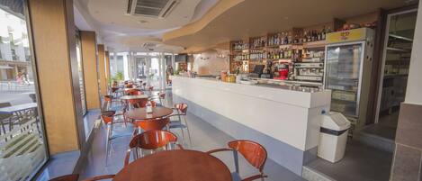 A restaurant with a long counter, multiple tables, and chairs. There are bottles and a refrigerator on the counter.