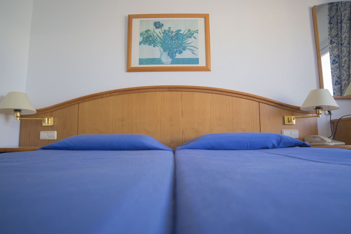 Una cama doble con ropa de cama azul, dos lámparas de noche, un cuadro en la pared y una mesita de noche con un teléfono.