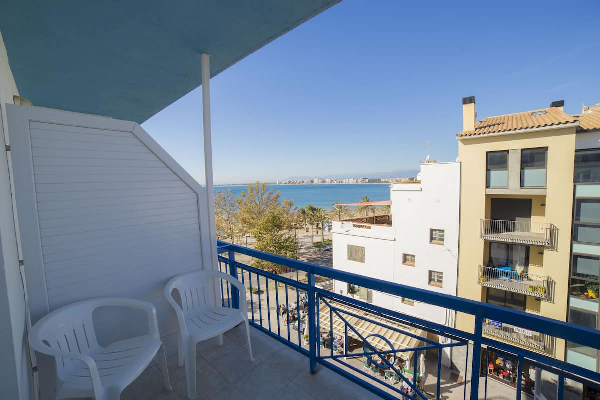 Un balcón con dos sillas de plástico blanco, barandilla azul y vista al mar y edificios.
