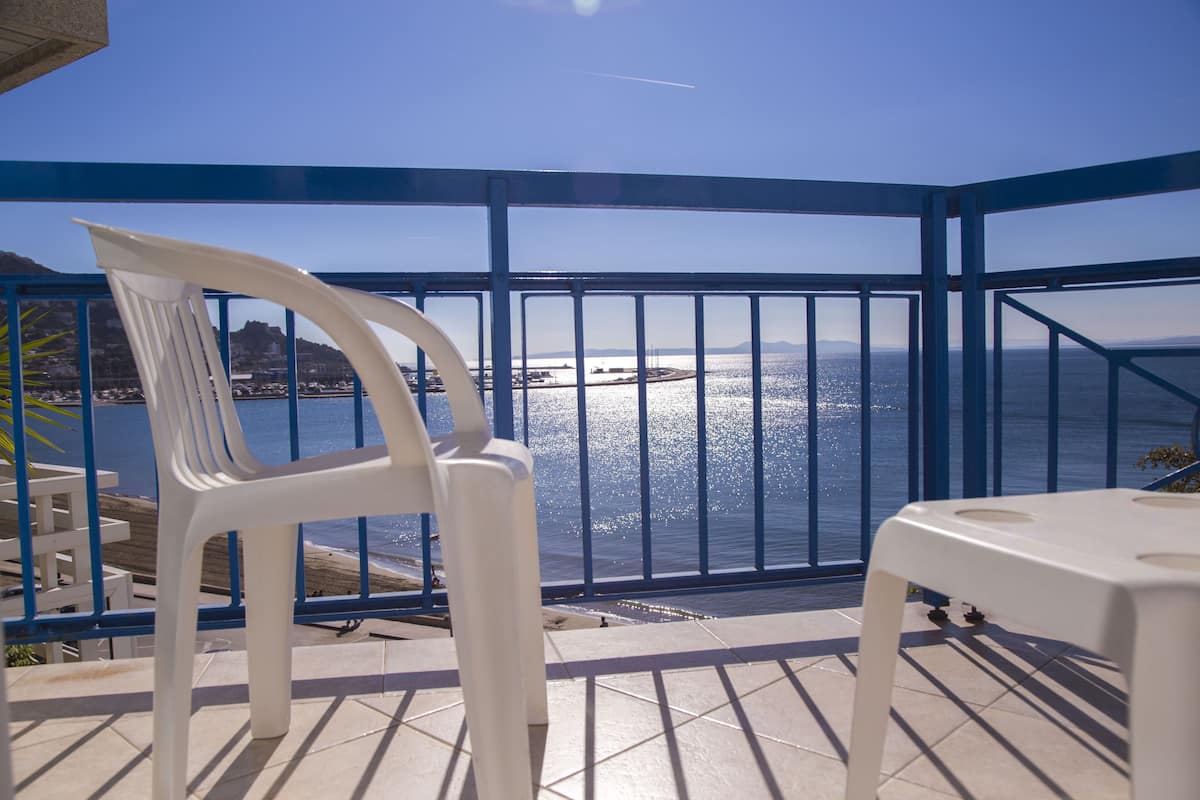 Un balcón con una silla y mesa de plástico blanco, con vista a una masa de agua, una costa lejana y montañas.