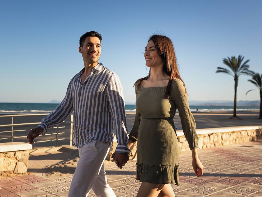 A couple walking hand in hand on a paved promenade with a beach and palm trees in the background.