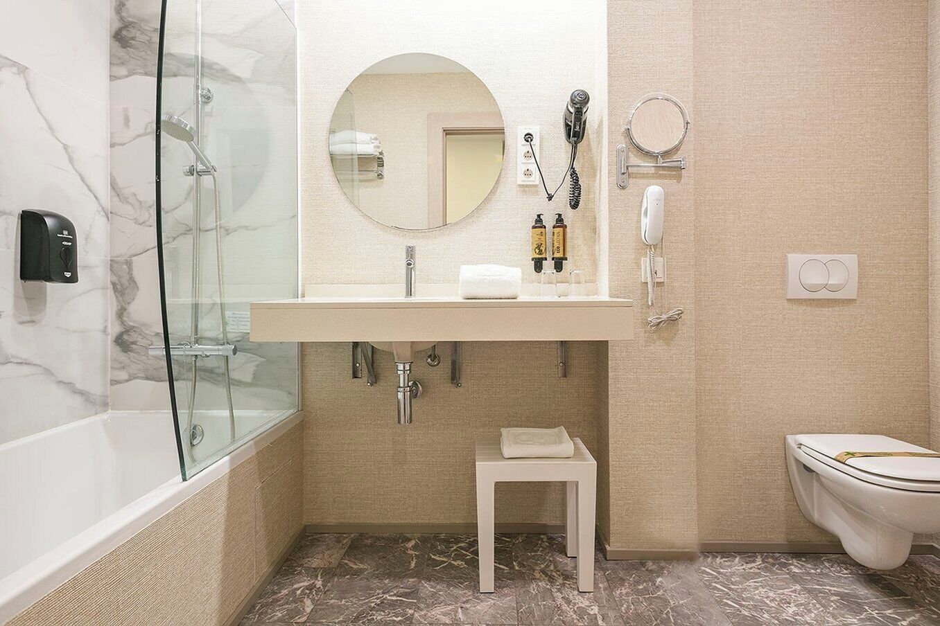 Une salle de bain avec une douche à parois de verre, un lavabo blanc avec un miroir, des toilettes et un petit tabouret.