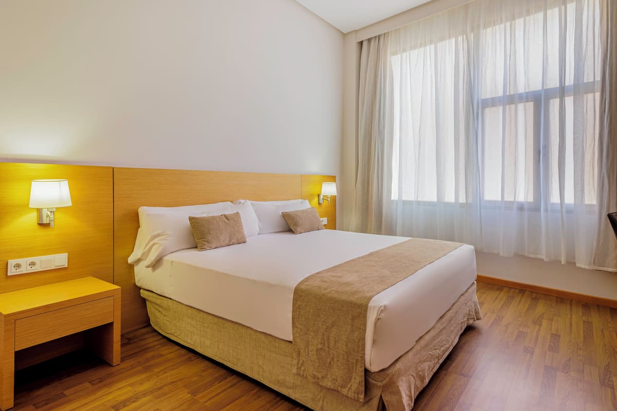 A hotel room with a bed, bedside lamps, a nightstand, and a window with sheer curtains.