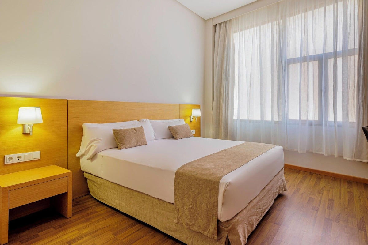 A hotel room with a large bed, bedside lamps, a wooden nightstand, and a window with sheer curtains.