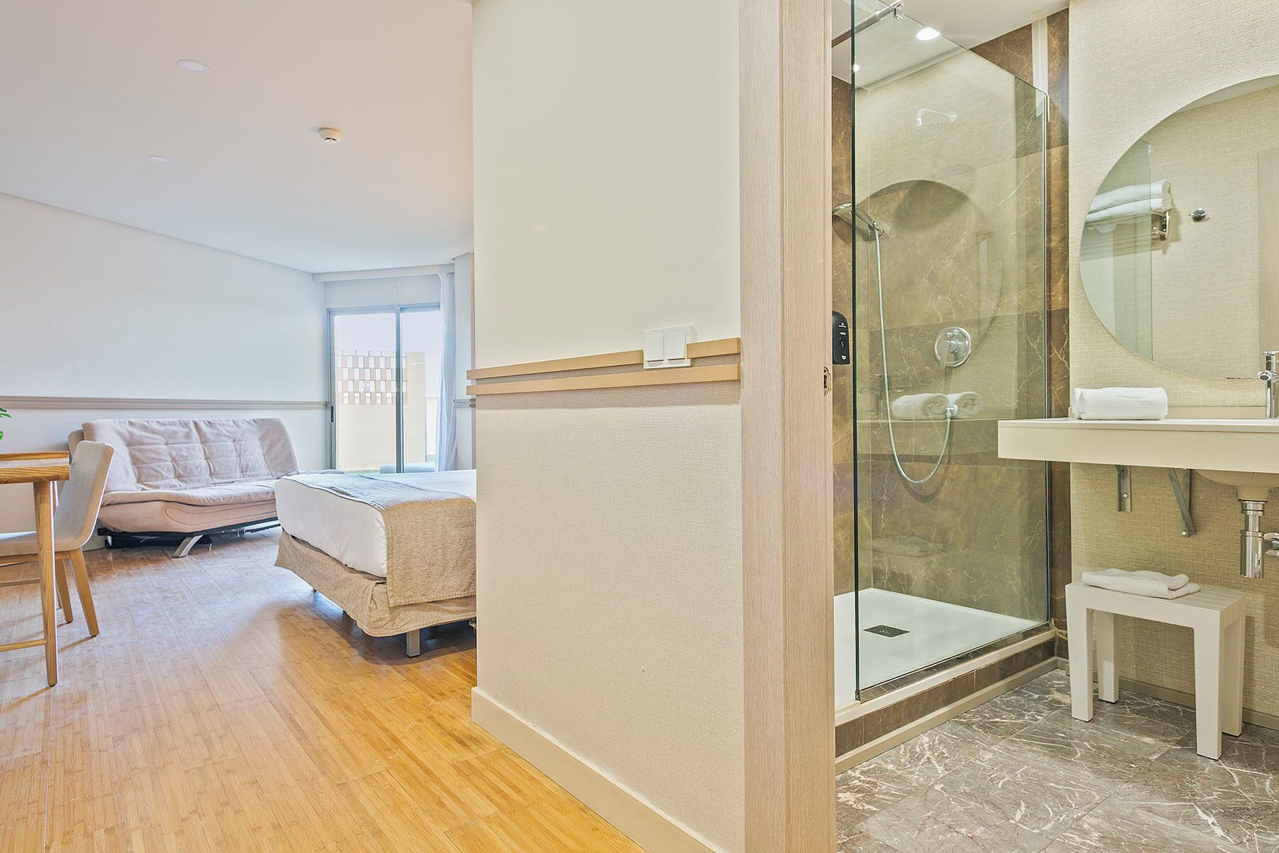 A hotel room with a bed, a sofa, a glass-enclosed shower, and a bathroom with a sink and mirror.