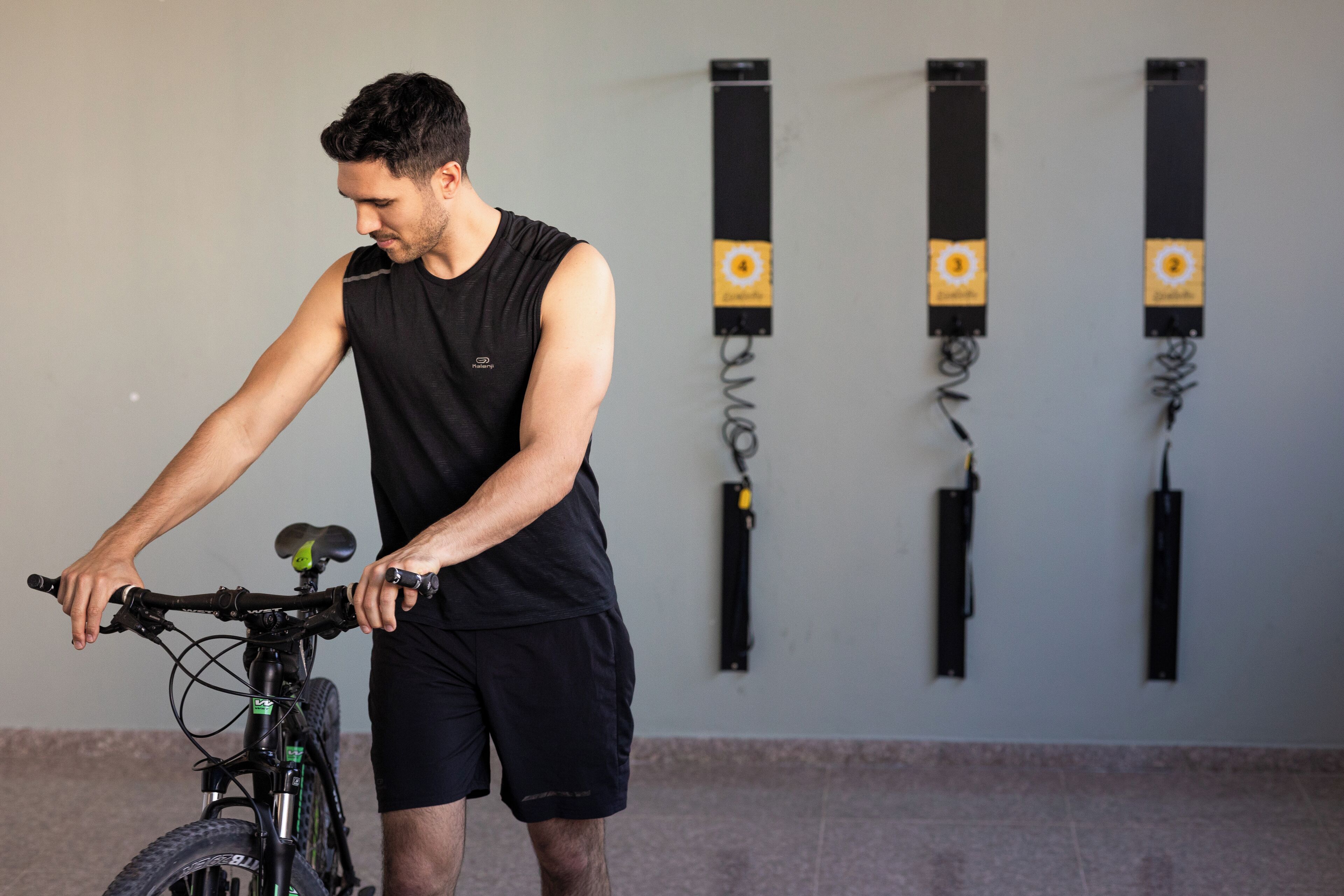 Una persona ajustando una bicicleta en una habitación con una pared lisa y tres máquinas de ejercicio negras.