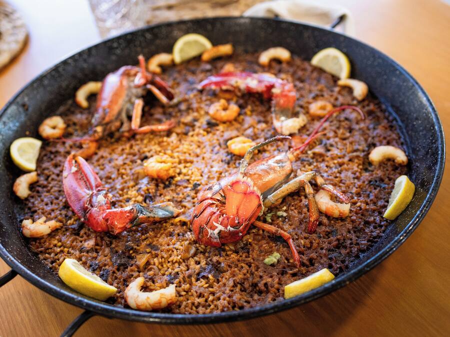 Una paellla de mariscos negra y abundante, adornada con rodajas de limón y camarones.
