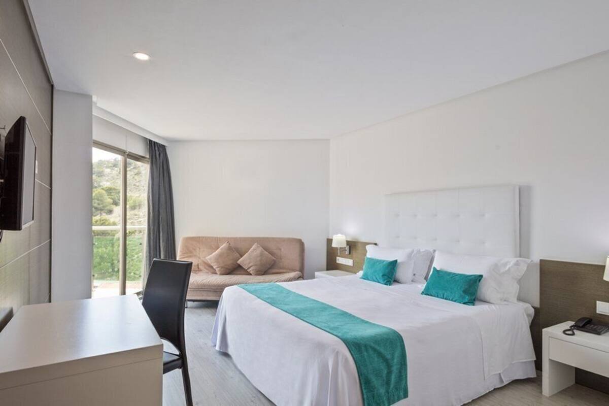 Habitación de hotel con cama, escritorio, silla, televisor y ventana con cortinas.