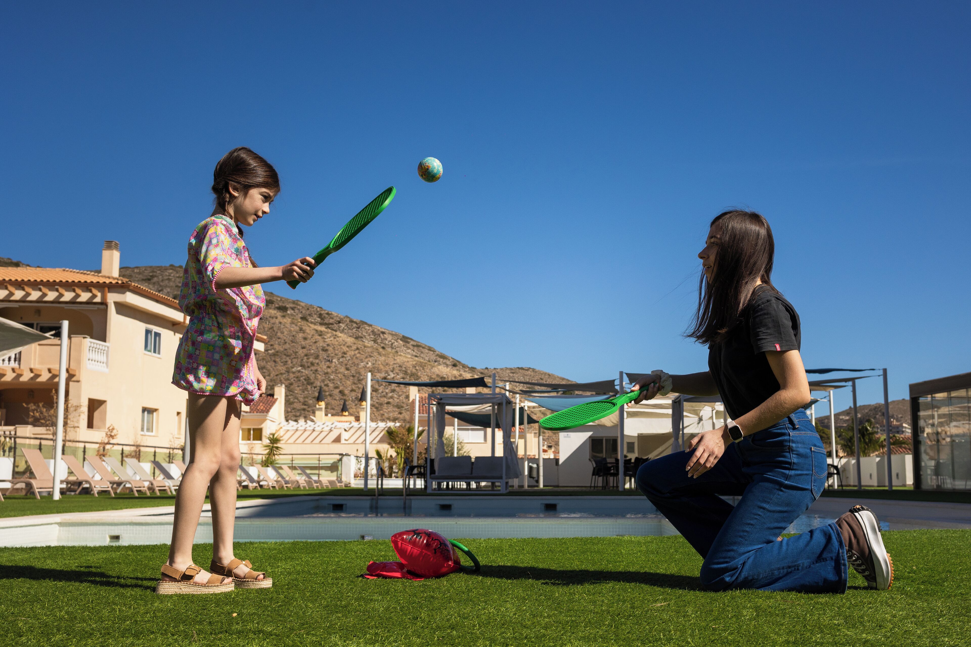 Una persona jugando con una pelota cerca de una piscina, con un edificio y colinas al fondo.