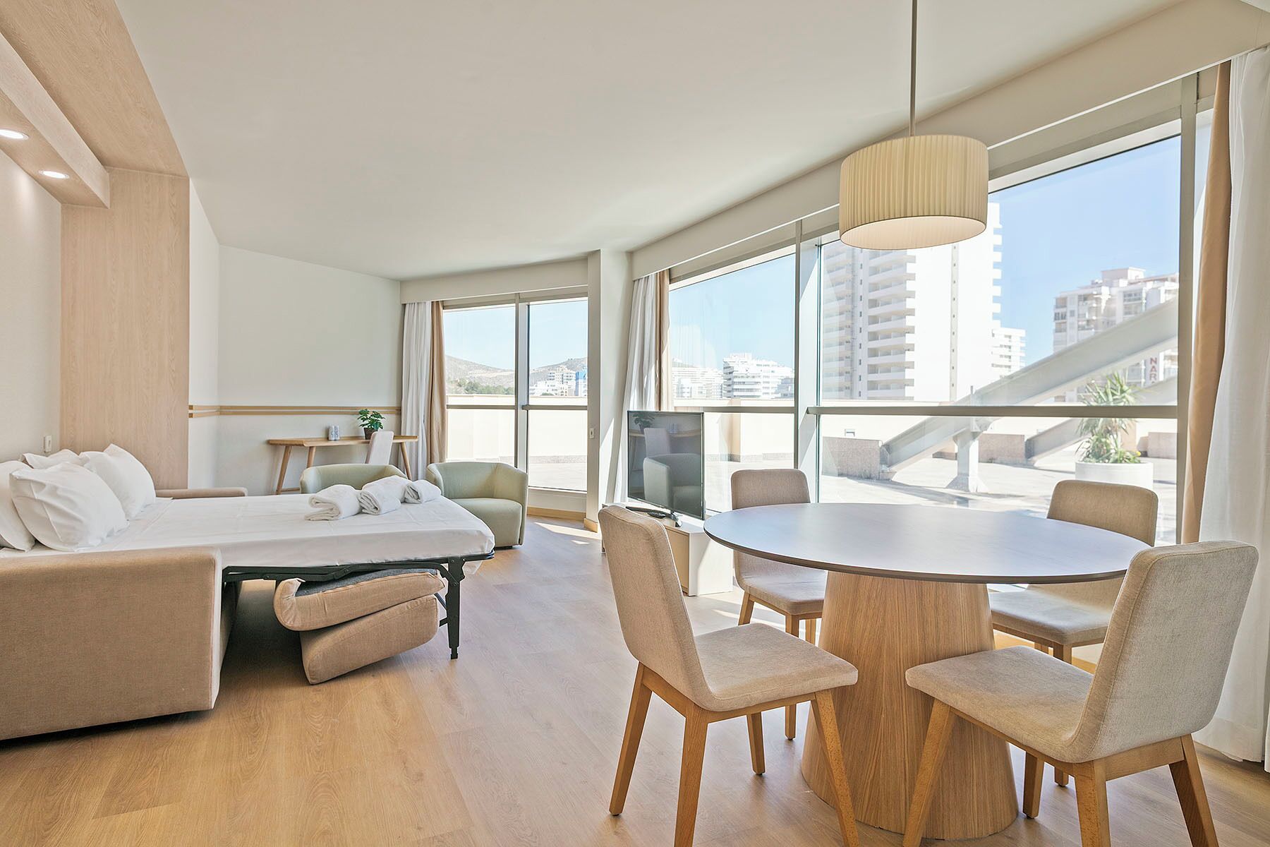 A modern hotel room with a bed, a sofa, a dining table with chairs, a TV, and large windows offering city views.