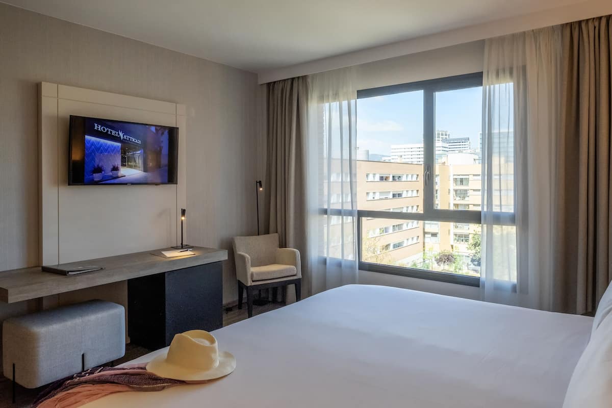 Une chambre d’hôtel avec un lit, un téléviseur fixé au mur, une chaise, un bureau et une grande fenêtre avec des rideaux.