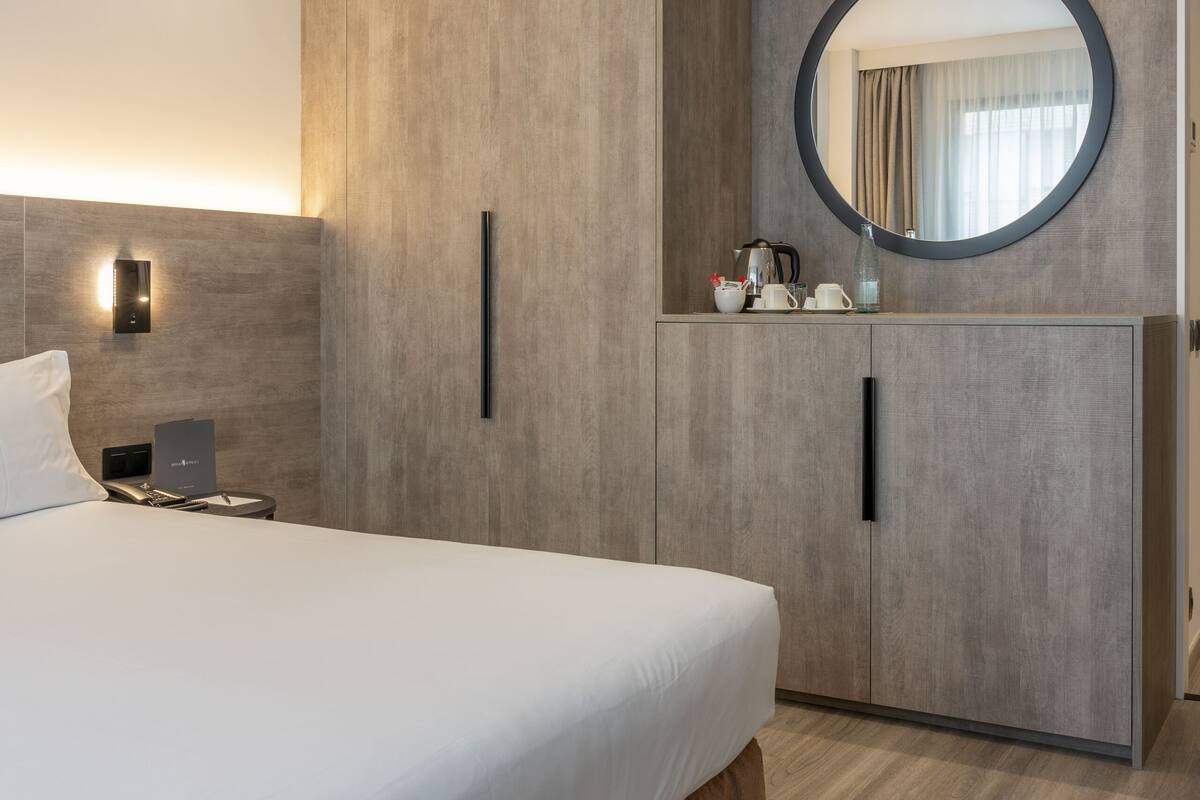 Une chambre d’hôtel moderne avec un grand lit, une armoire, un miroir rond et une table de chevet avec une lampe.