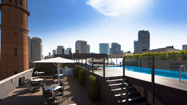 Un espace piscine aménagé sur le toit, avec des chaises longues, des tables et une vue panoramique sur la ville en arrière-plan.