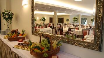 Un salón de banquetes con mesas dispuestas para un evento formal, un espejo grande y ornamentado que refleja la sala, y una mesa de buffet con frutas y pasteles.