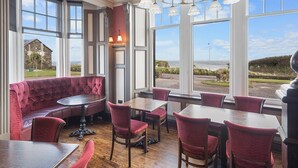 A restaurant with a red upholstered bench, wooden tables, and chairs. Large windows offer a view of the sea and a stone building. - The Ugadale Hotel and Cottages (Campbeltown)