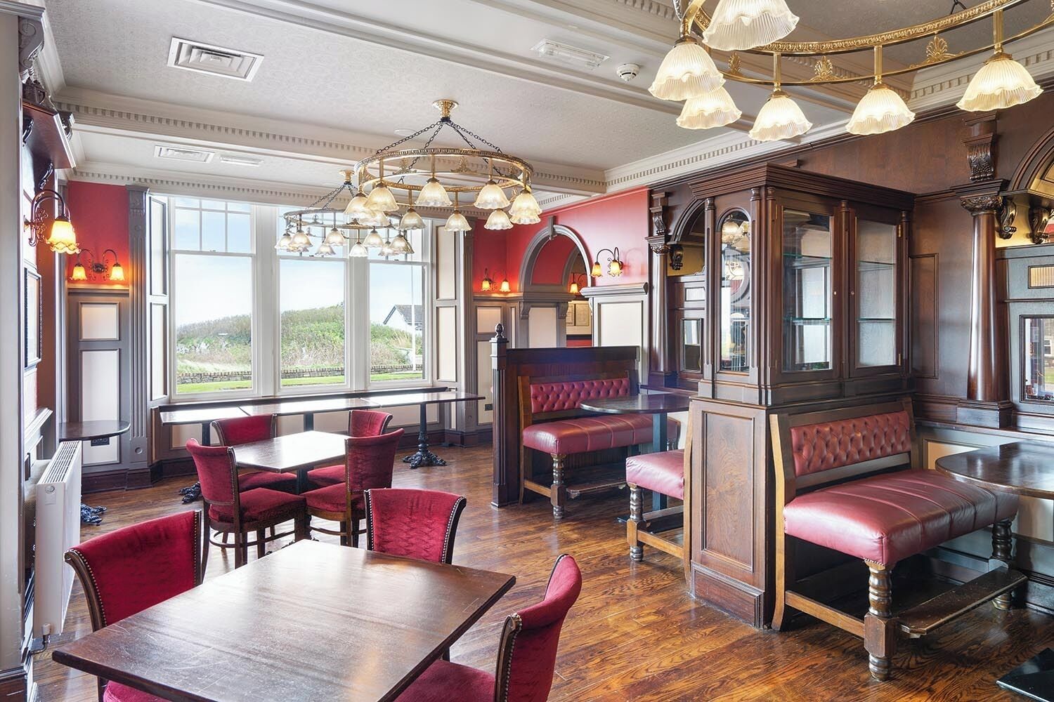 A restaurant with wooden tables and red upholstered booths, large windows, and ornate chandeliers.